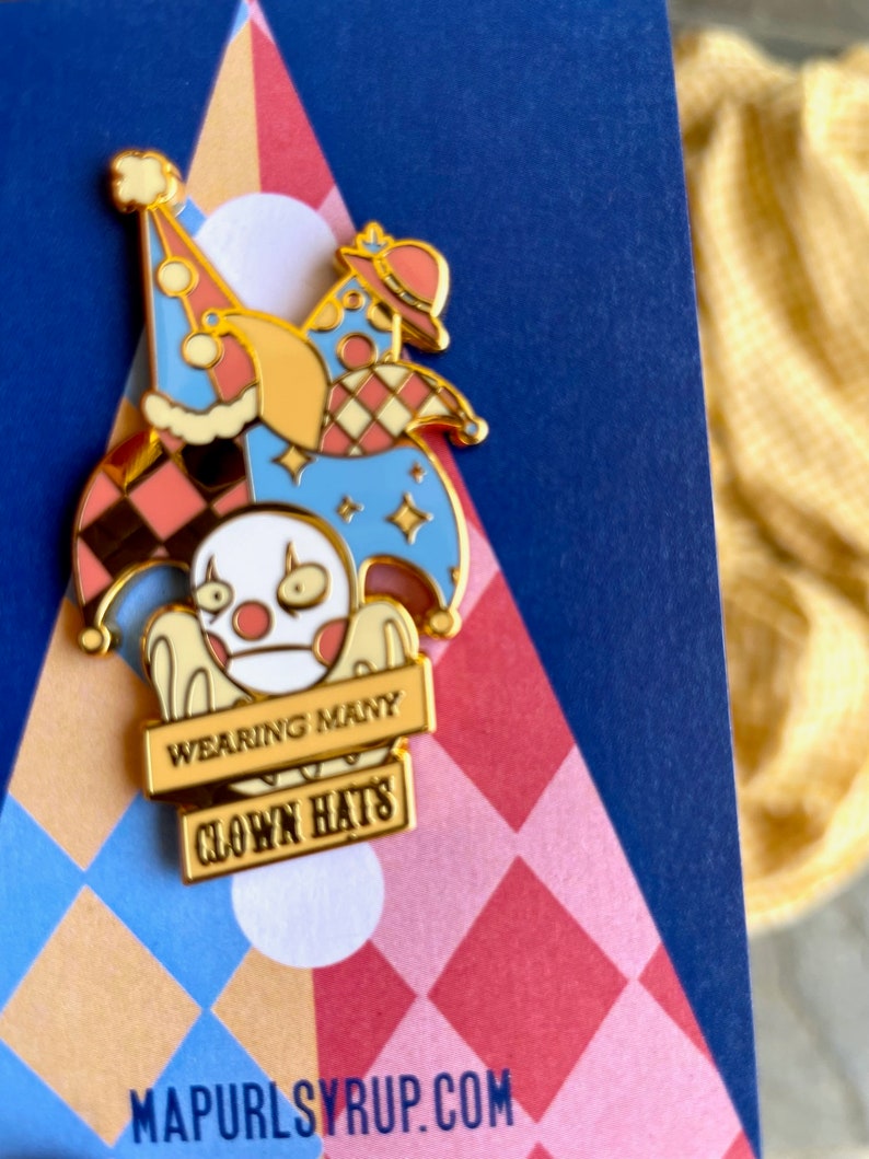 May include: Gold enamel pin featuring a white cartoon character wearing multiple colorful hats. The character is holding a sign that reads "Wearing Many Clown Hats".