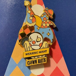 May include: A gold-colored enamel pin featuring a cartoon clown wearing multiple hats. The hats are a jester's hat, a party hat, and a top hat. The pin has a banner that reads "Wearing Many Clown Hats".