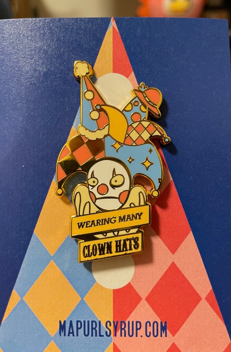 May include: A gold-colored enamel pin featuring a cartoon clown wearing multiple hats. The hats are a jester's hat, a party hat, and a top hat. The pin has a banner that reads "Wearing Many Clown Hats".