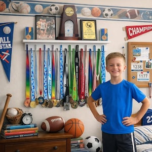May include: A boy stands in front of a wall decorated with sports-themed items. A shelf holds trophies and framed awards. Below, a row of medals hangs. A football, basketball, and soccer ball sit on a wooden nightstand, along with a baseball and a baseball bat.