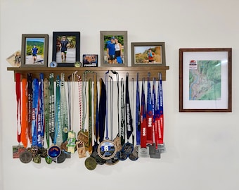 May include: A wooden shelf with a collection of medals hanging on hooks. The medals are mostly blue, red, and green with various logos and text. There are also framed photos of people running and a map of a trail.