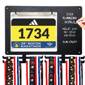 May include: Black metal medal display rack with a clear plastic holder for a race bib. The bib is yellow with black text and shows the number 1734, the 128th Boston Marathon, and the Bank of America logo. The rack has three rows of hooks for hanging medals. A chalkboard section on the right side of the rack has the text "2023 Running Goals" and handwritten goals for a mile, 5k, and 10k run.
