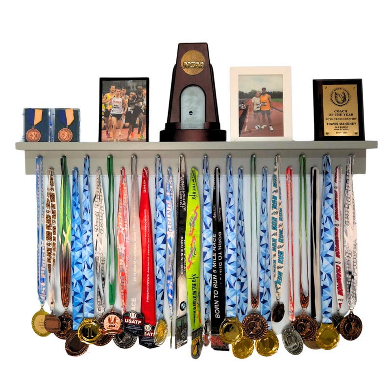 Pu&ograve; includere: Una mensola grigia in legno che espone medaglie di corsa, trofei e foto incorniciate. Le medaglie hanno nastri colorati e vari disegni. La mensola contiene anche un trofeo con il logo NCAA e una targa con scritto "Allenatore dell'anno".