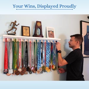 Pu&ograve; includere: Una mensola bianca che espone medaglie con nastri colorati. Le medaglie sono di varie dimensioni e colori, tra cui oro, argento e bronzo. Sopra le medaglie ci sono trofei e una foto incorniciata. Il testo "Your Wins, Displayed Proudly" &egrave; in alto.
