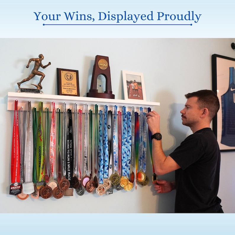 Pu&ograve; includere: Una mensola bianca che espone medaglie con nastri colorati. Le medaglie sono di varie dimensioni e colori, tra cui oro, argento e bronzo. Sopra le medaglie ci sono trofei e una foto incorniciata. Il testo "Your Wins, Displayed Proudly" &egrave; in alto.