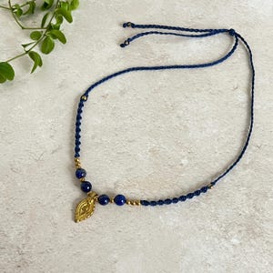 May include: A handmade navy blue macramé necklace with gold-coloured accents and a gold pendant. The necklace features dark blue beads and a braided cord. The pendant has an intricate leaf design.