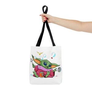 May include: A white tote bag with black straps features a cartoon illustration of a green character with large eyes and pink clothing. The character is holding a wand and surrounded by bubbles. The text "Dood" is visible on a can in the image.