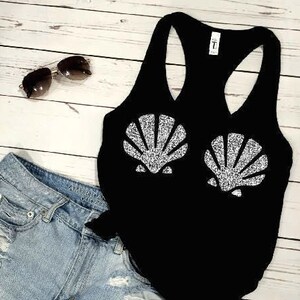 May include: A black tank top with two glittery seashell designs, paired with light-wash denim shorts and sunglasses. The shorts have distressed detailing. The text "PIERCED N PRETTY" is visible at the bottom.
