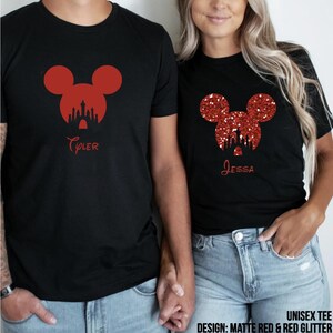 May include: Two black t-shirts featuring red glitter Mickey Mouse ears and a silhouette of Cinderella's castle. The shirts are personalised with the names "Tyler" and "Jessa". The design is described as "Matte Red & Red Glitter".