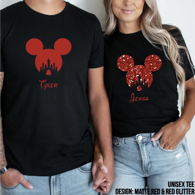 May include: Two black t-shirts featuring red glitter Mickey Mouse ears and a silhouette of Cinderella's castle. The shirts are personalised with the names "Tyler" and "Jessa". The design is described as "Matte Red & Red Glitter".