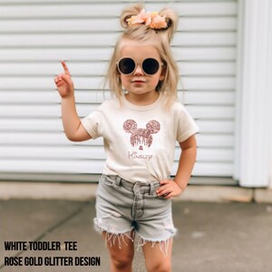 May include: White toddler tee shirt with a rose gold glitter Mickey Mouse design and the name "Kinsley" written underneath.