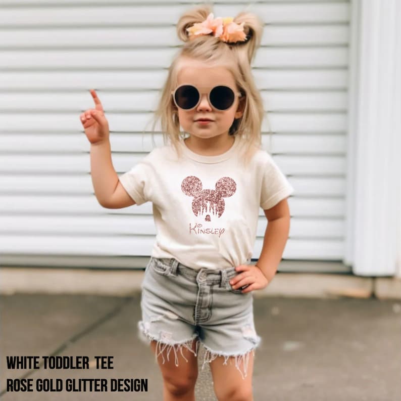 May include: White toddler tee shirt with a rose gold glitter Mickey Mouse design and the name "Kinsley" written underneath.