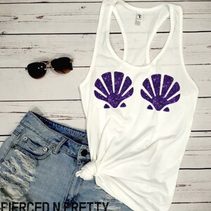 May include: White tank top with a racerback design, featuring two purple glitter seashell designs. The image also includes denim shorts, sunglasses, and the text "PIERCED N PRETTY" at the bottom.