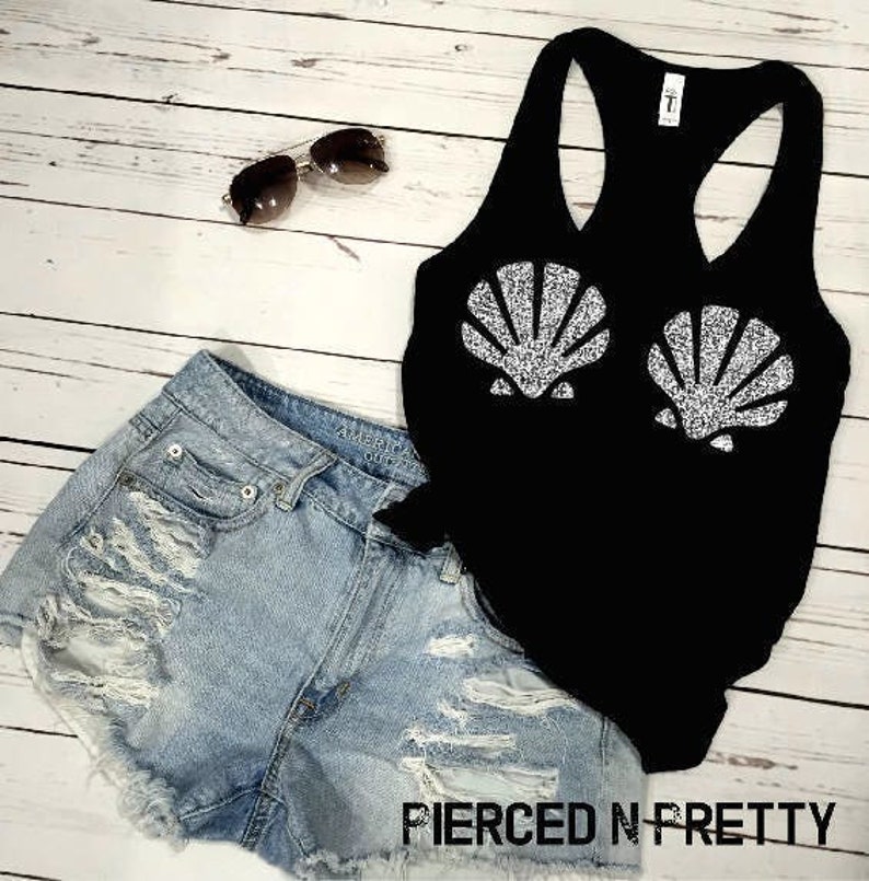 May include: A black tank top with two glittery seashell designs, paired with light-wash denim shorts and sunglasses. The shorts have distressed detailing. The text "PIERCED N PRETTY" is visible at the bottom.