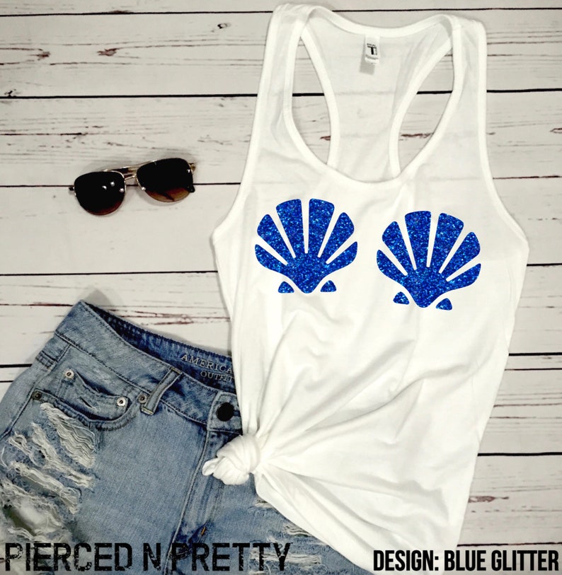 May include: White tank top with a racerback design. The shirt features two blue glitter seashell designs on the front. Sunglasses and denim shorts are also visible. The text "Pierced N Pretty" and "Design: Blue Glitter" are printed on the bottom.