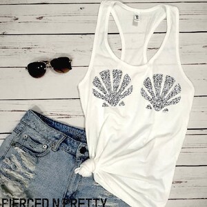 May include: White tank top with a seashell design in silver glitter. The top is paired with denim shorts and sunglasses. The text "PIERCED N PRETTY" is visible at the bottom of the image. The outfit is laid out on a light wood background.