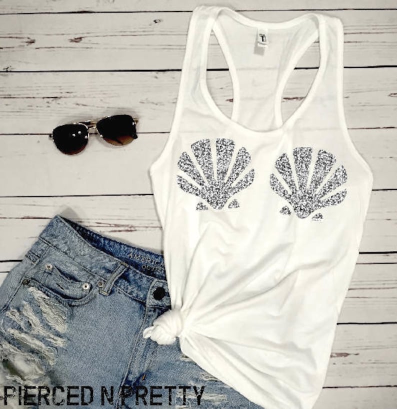May include: White tank top with a seashell design in silver glitter. The top is paired with denim shorts and sunglasses. The text "PIERCED N PRETTY" is visible at the bottom of the image. The outfit is laid out on a light wood background.