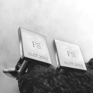 Może przedstawiać: Para srebrnych spinek do mankiet&oacute;w z wygrawerowanymi inicjałami "FS" i datą "26.07.2003".