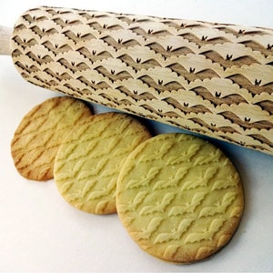 May include: A wooden rolling pin with a bat pattern is displayed next to three round cookies with the same bat design. The rolling pin and cookies are light brown, creating a cohesive theme. The cookies are approximately 3 inches in diameter.