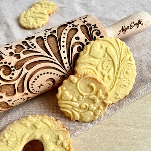 Può includere: Un mattarello di legno con un motivo floreale intagliato e il testo "Algio Crafts" sul manico. Il mattarello viene utilizzato per creare motivi decorativi sui biscotti. Nell'immagine sono mostrati tre biscotti con lo stesso motivo floreale.