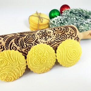 Può includere: Un mattarello di legno con un design inciso a fiocchi di neve e volute, insieme a tre biscotti in rilievo. I biscotti e il mattarello sono marrone chiaro. Il mattarello è lungo circa 38 cm.