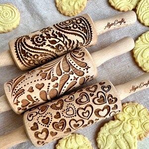 May include: Three wooden rolling pins with different engraved designs. The designs include paisley, floral, and hearts. The rolling pins are all made by Algis Crafts.