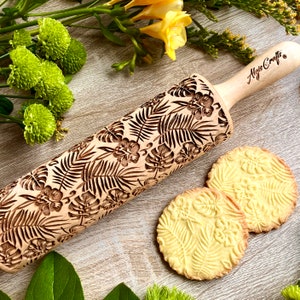 Könnte beinhalten: Ein Holz-Nudelholz mit einem tropischen Blatt- und Blumendesign. Das Nudelholz ist mit dem Text "Algis Crafts" graviert. Zwei Kekse mit dem gleichen Design sind auf dem Bild zu sehen.