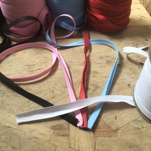 May include: Assortment of colorful bias tape in pink, black, red, blue, and white. The tape is rolled up on spools and laid out on a wooden surface.