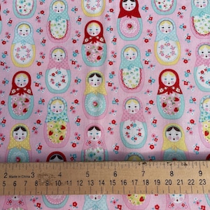 May include: Pink fabric with a repeating pattern of nesting dolls. The dolls are wearing colourful dresses and have smiling faces. The fabric is decorated with small pink flowers.