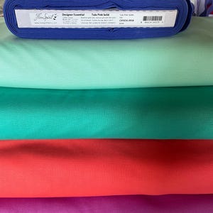 May include: A stack of colorful fabric bolts in shades of blue, mint green, teal, red-orange, and purple. The top bolt is a deep blue with a label that reads "Free Spirit Designer Essential Tula Pink Solid". The fabric is tightly rolled and stacked.