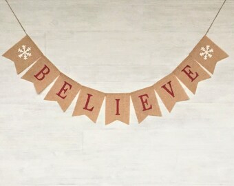 May include: A burlap banner with the word "BELIEVE" in red letters and snowflakes on each end. The banner is hanging against a white wood background.