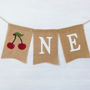 May include: A burlap banner with the word "ONE" spelled out in white paint. The first letter is decorated with a red cherry with a green stem and leaf.