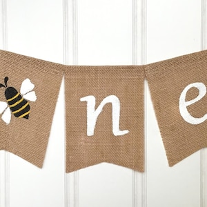 May include: A burlap banner with the word "one" spelled out in white paint. The first "o" is replaced with a black and yellow bee with white wings.