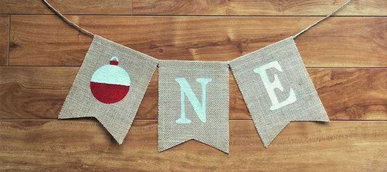Peut inclure: Une banni&egrave;re en toile de jute avec le mot "ONE" en lettres bleu clair et un flotteur de p&ecirc;che rouge et blanc sur le c&ocirc;t&eacute; gauche. La banni&egrave;re est enfil&eacute;e sur une ficelle et est parfaite pour une f&ecirc;te de premier anniversaire.