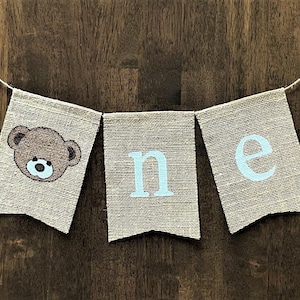 May include: A burlap banner with the word "one" in light blue letters and a brown teddy bear face on the first flag.