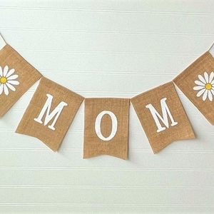 May include: A burlap banner with the word "MOM" in white letters and a daisy on each end. The banner is strung on a white string.