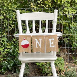 May include: A white wooden high chair featuring a three-piece burlap banner hanging from the tray. The banner displays a red and white fishing bobber graphic on the first panel and the white letters O, N, and E on the subsequent panels.