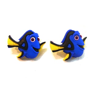 May include: A pair of blue fish-shaped earrings featuring a smiling face, black accents, yellow fins, and pink eyes. These earrings are inspired by the character Dory from the animated film.