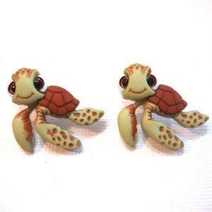 May include: Two turtle-shaped embellishments. Each turtle has a light green body with brown spots and a reddish-brown shell. They have large, dark eyes and are smiling. The turtles are likely made of plastic or resin.