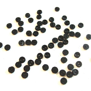May include: A scattering of small, round, black buttons with two holes each, arranged on a white surface. These buttons are likely for sewing or crafting projects, offering a classic and versatile design element for various apparel and craft items.