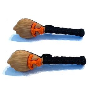 May include: Two miniature broom charms. Each charm features a light brown broom head, an orange accent, and a black handle. These charms are likely for Halloween or witch-themed crafts.