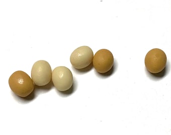 May include: Six small, oval-shaped objects in varying shades of brown and off-white are scattered on a white surface. The objects resemble miniature eggs, with smooth, glossy surfaces. The arrangement is casual, with the objects spaced apart.