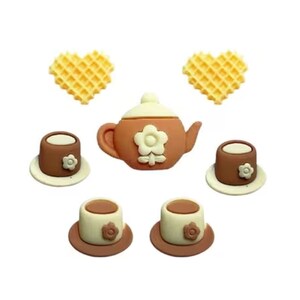 May include: A collection of miniature ceramic tea party decorations. The set includes a brown teapot with a white flower, matching teacups, saucers, and heart-shaped waffles. The pieces are arranged on a white background.