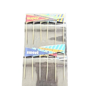 May include: A package of decorative cake toppers. Each topper has a wooden stick with a colorful banner. One banner reads "super" in red, and the other reads "sweet" in blue. The banners have geometric patterns.