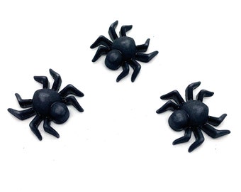May include: Three small, black plastic spiders are arranged on a white background. Each spider has a rounded body and eight legs, with a slightly glossy finish. The spiders are likely decorative items.