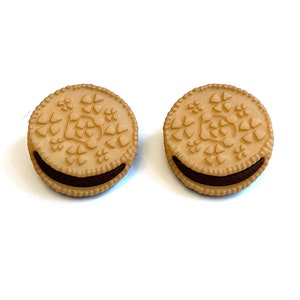 May include: Two tan-colored, round earrings resembling chocolate sandwich cookies. Each earring has a detailed, embossed design on the top and a dark brown filling. The earrings are set against a white background.