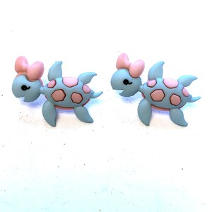 May include: Two turtle-shaped embellishments. Each turtle is light blue with a pink shell pattern and a pink bow. The turtles have black eyes and are facing each other. These are likely used for crafts or jewelry.