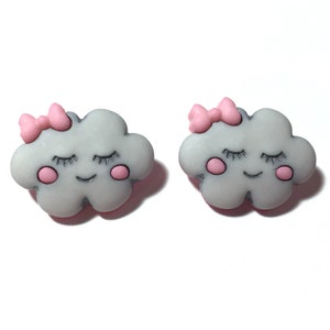 May include: Two gray cloud-shaped accessories with pink bows and rosy cheeks. Each cloud has closed eyes and a smiling mouth. The accessories are likely earrings or decorative embellishments.