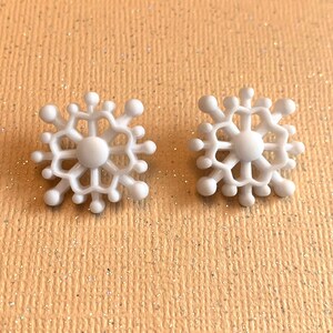 May include: Two white snowflake-shaped stud earrings. Each earring features a central circular design with radiating arms and small, rounded accents. The earrings are set against a light orange background with a glittery texture, suggesting a winter or holiday theme.