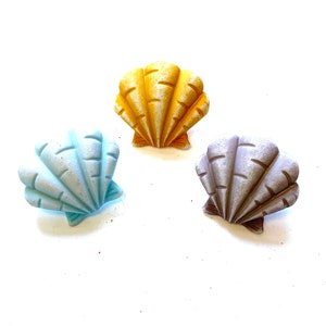 May include: Three seashell-shaped decorative objects in shades of yellow, blue, and gray. Each shell has a textured surface with detailed ridges. The shells are arranged on a white background.